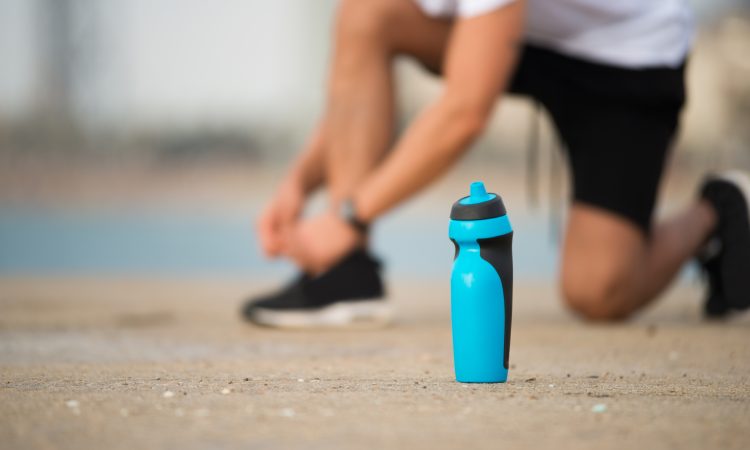 Blue fitness shaker bottle placed on ground. Sportsman tying shoelace in background. Close-up of bottle with drinking water or protein. Restoring strength concept
