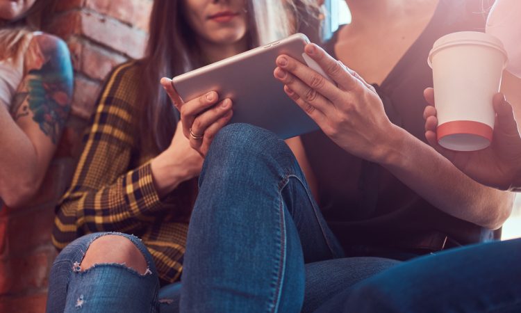 Group of female friends in casual clothes discussing while looking something on a digital tablet in a room with a loft interior.