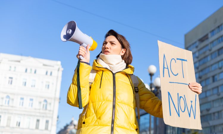 Portrait of young woman standing outdoors in town and showing table Act Now. Female demonstrating board with protest against pandemic, political or environmental issues. Single protest.