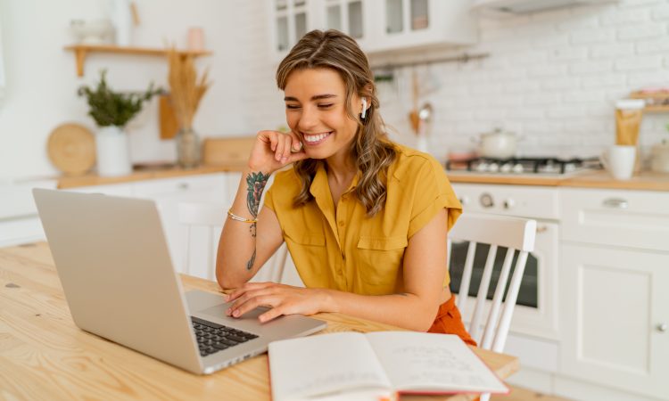 Pretty miling student woman using laptop and writing notes on her modern light kitchen.