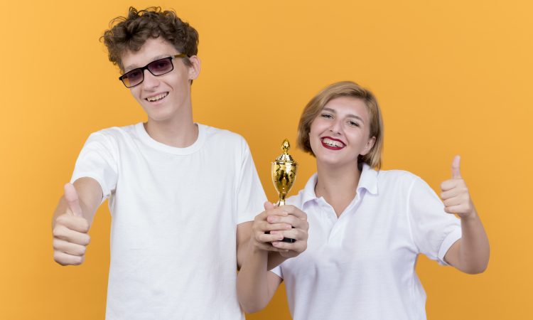 young sporty couple man and woman standing together holding trophy looking at camera showing thumbs up smiling standing over orange background