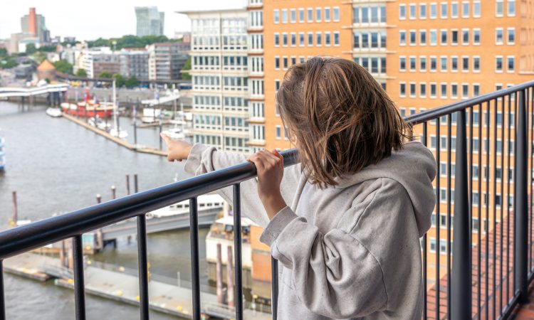 A child in a hoodie stands on a high-rise balcony, pointing toward the river harbor with boats. The scene reflects curiosity and urban life.