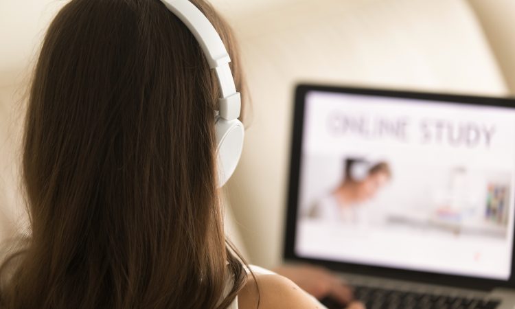 Woman in headphones sitting on sofa with laptop on knees studying online, using web services to learn languages. Female student proceeds educational course in Internet. Close up view over the shoulder