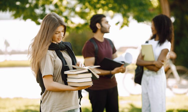 Focus on a caucasian girl who standing sepately. Group of international students standing together in park at university. Blurred african girl and indian boy talking outdoors.